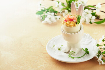 Stuffed or deviled eggs with yolk, shrimp, pea microgreens with paprika in rabbit-shaped stand for easter table decorate fresh cherry or apple blossoms on light background. Traditional dish for Easter
