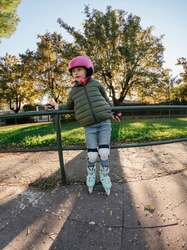 A child in a pink helmet and green jacket is rollerblading in a park, with trees and a sunset in the background.