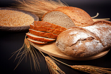 Whole and sliced breads, wheat ears and wheat on wooden plate on black background. Photograph.