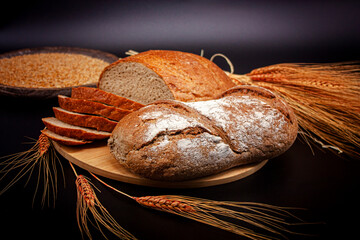 Whole and sliced breads, wheat ears and wheat on wooden plate on black background. Photograph.