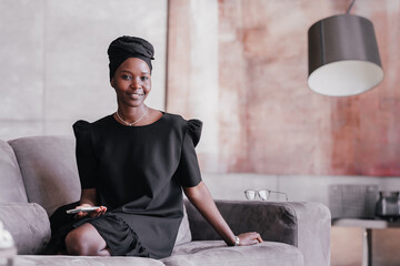 satisfied African young woman in a black dress in a black tulip is sitting on the couch at home...