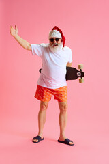 Cool Santa Claus contemporary dressed for summer with style sunglasses holding skateboard against pastel pink studio background.