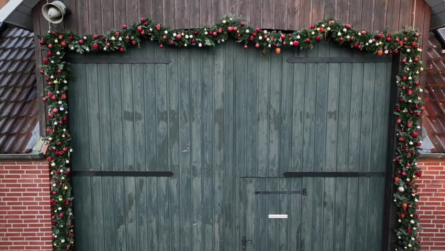 Aerial footage of light emerald barn door decorated with Christmas garlands and sunrise reflection in the window and solar panels on the barn roof. Barn with holiday decorations like stars, balls.