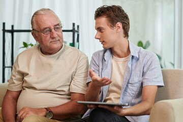 Positive young man explaining grandfather how to install apps on tablet computer