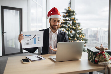 Confident adult pointing at graphic materials while giving online presentation from office on background of Christmas tree. Focused worker providing statistical analysis using modern technologies.