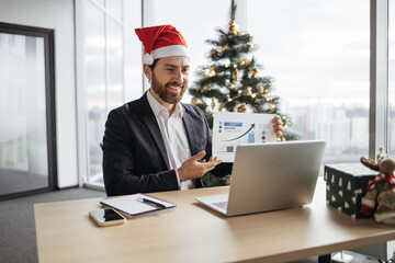 Confident adult pointing at graphic materials while giving online presentation from office on background of Christmas tree. Focused worker providing statistical analysis using modern technologies.