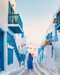 Mykonos Greece, Little Venice Mykonos Greece, Asian woman in a blue dress walking at the street of sunset at ocean fron at Mykonos town Europe - obrazy, fototapety, plakaty