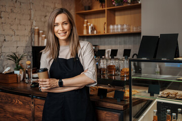 A portrait of a poised female barista holding a takeaway coffee cup, standing in her modern cafe