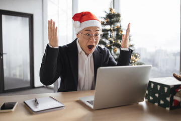 Emotional bespectacled adult in Santa hat lifting hands in excitement while looking at computer screen in office decorated with Christmas tree. Delighted asian male making successful holliday purchase