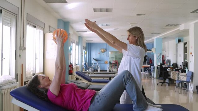 Blonde physiotherapist is showing to her patient how to do some exercises with a ball for recover her injury on her shoulders