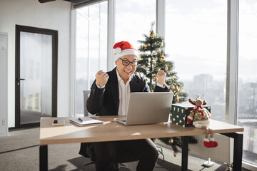 Emotional adult in Santa hat lifting fists of hands in excitement while looking at computer screen in office decorated with Christmas tree. Delighted asian male making successful holliday purchase