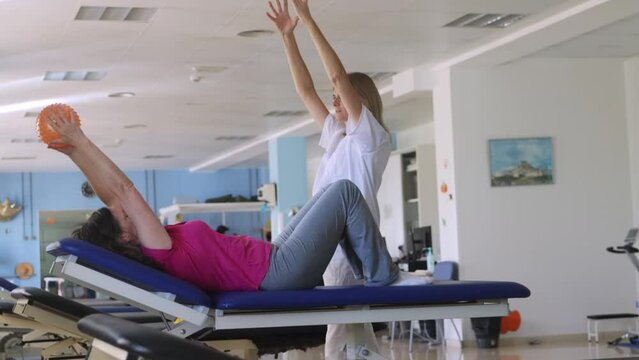 Handheld clip of a woman patient doing an exercise with a ball at the rehabilitation center. The woman physiotherapist is in front of her teaching it.