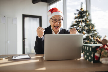 Emotional adult in Santa hat lifting fists of hands in excitement while looking at computer screen in office decorated with Christmas tree. Delighted asian male making successful holliday purchase