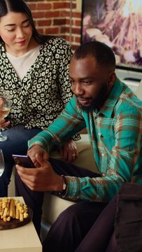 Vertical video Inclusive group of friends at house party using smartphone to laugh at social media posts made by acquaintance. Amused guests in living room using phone to look at cringe images posted