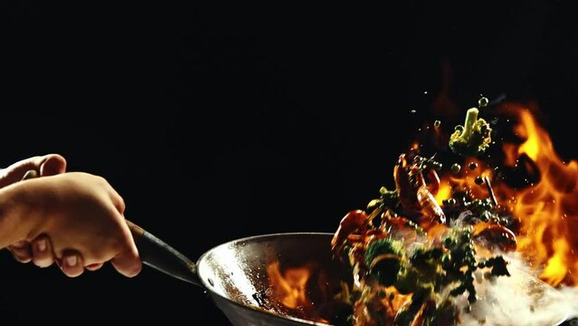 Chef cooking delicious wok noodles, frying vegetables and shrimps in pan with fire flames against black background. Concept of Asian food, cuisine, restaurant, taste, cooking, recipe