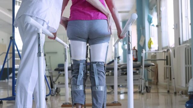Rear view of an unrecognizable woman with prosthesis at both legs learning to walk another time at the parallel bars at the hospital. Her nurse is at her side helping to her.