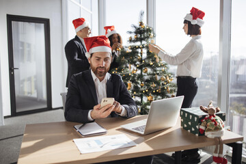Businessman in Santa hat sitting at table and using modern smartphone in decorated festive office....