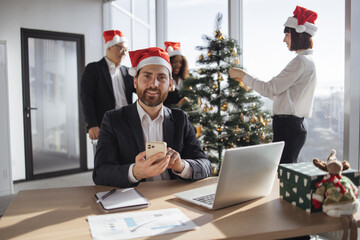 Businessman in Santa hat sit at table and using smartphone in decorated festive office looking at camera. Male freelancer taking break during working process while coworkers decorating Christmas tree