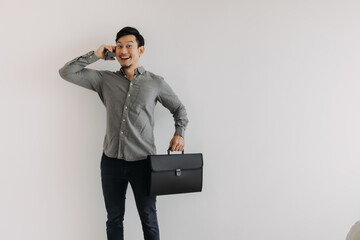 Asian Thai business man happy face using mobile phone and holding black file folder bag while standing and looking at camera isolated over white background wall.