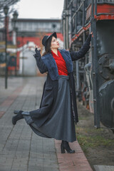A beautiful girl in a black coat and hat on the running board of an old steam locomotive. A young woman with long dark hair. Vintage portrait of the last century, retro journey.