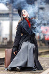 A beautiful girl in a black coat and hat with a suitcase in her hands near an old steam locomotive. A young woman with long dark hair. Vintage portrait of the last century, retro journey.