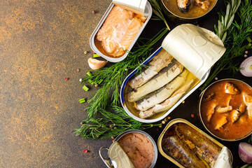 Different open tin cans with canned fish among spices and herbs on a brown background, canned salmon and mackerel, sprat and sardine, tuna and herring and fish pate, top view