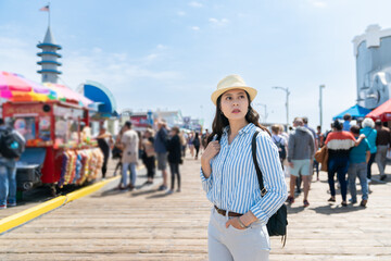 stylish asian Japanese girl backpacker with hand in pocket exploring busy santa monica pier near...