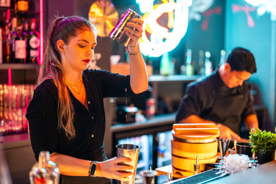 Side view of Confident barkeeper with blonde hair standing at counter while preparing beverage in cocktail shaker in bar