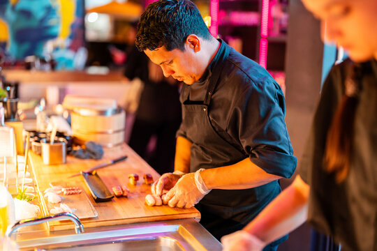 Focused Male cook in black uniform and gloves preparing fresh sushi on wooden chopping board at kitchen in nightclub