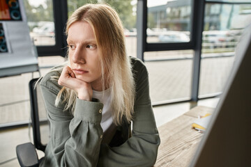 pensive young androgynous person in green shirt sitting at desk with hand under chin, business