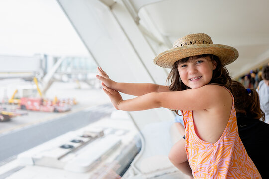 Side view of faceless girl traveler wearing straw hat and casuals looking at camera while contemplating airplane on taxiway through window at airport