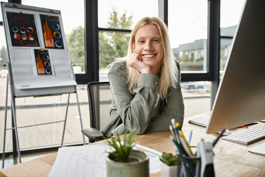 joyful attractive non binary person smiling at camera with hand under chin, business concept