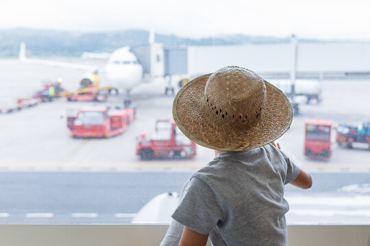 Back view of faceless boy traveler wearing straw hat and casuals looking at passenger airplane on taxiway through window at airport