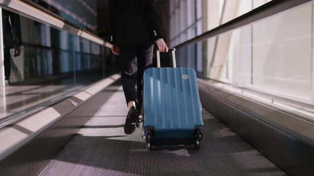 Caucasian young woman in black business suit confidently pulling a blue suitcase on an airport moving walkway, symbolizing efficiency and pace of modern business travel