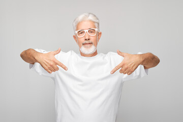 Gray-haired mature man 60 years old in white T-shirt pointing finger down, demonstrating empty...