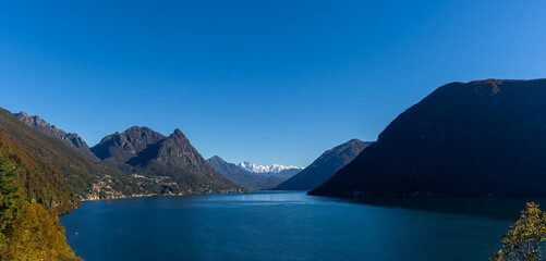 Fototapeta premium panorama landscape view of Lake Lugano with green hills and snow-capped mountain peaks under a blue sky