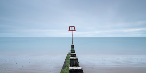 Long exposure of the sea with a groyne in centre that has a red shaped sign at the end