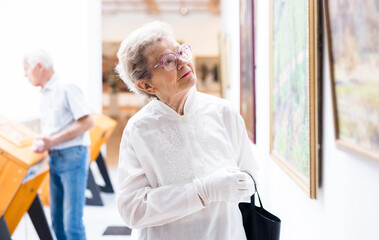 Mature woman examines paintings in an exhibition in hall of an art museum