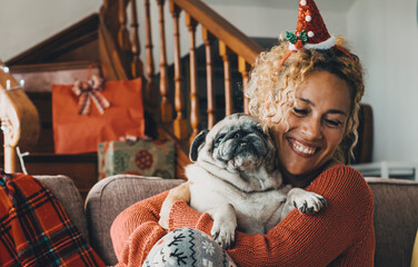 Dog owner female enjoy holiday christmas celebration at home alone with her pug best friend. Woman hugging and enjoying her dog wearing santa clasu hat and smiling a lot. Concept of friendship