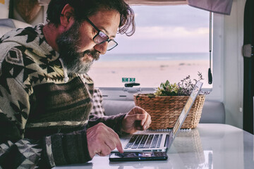 Working off grid in alternative office and online job lifestyle concept man. Adult people work on a laptop inside a camper van motor home. Smart working in travel vehicle transport. Using notebook - obrazy, fototapety, plakaty