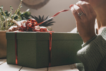 Close up of a woman unpacking a surprise gift box. Christmas or birthday anniversary present. Female people celebrate at home with a package and red tape. Green color mood image. Sharing gift moment - obrazy, fototapety, plakaty