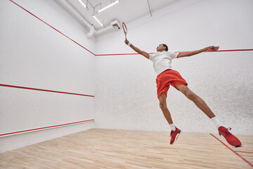 motion shot, energetic african american sportsman with racquet jumping and playing squash in court