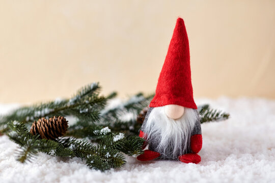 christmas, decoration and winter holidays concept - close up of gnome toy and fir branch with cone on artificial snow over beige background