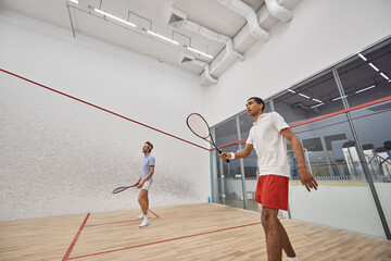 young and interracial men in sportswear playing squash inside of court, challenge and motivation
