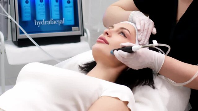 Cosmetology clinic. Professional female cosmetologist doing hydrafacial procedure while being a work. Attractive nice woman lying on the medical bed while having beauty procedures