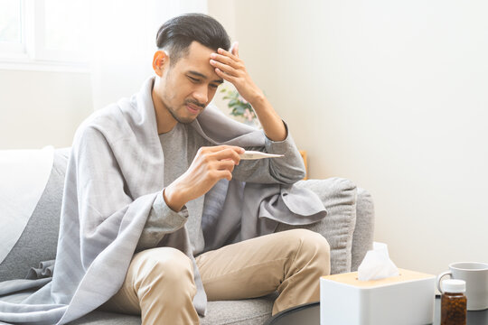 Sick, influenza asian young man, male have fever hand holding thermometer, touching forehead for check measure body temperature, illness while sitting rest on sofa at home. Health care on virus person