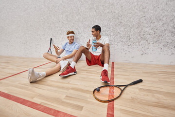 cheerful interracial sportsmen with squash racquets and bottle with water sitting on floor in court