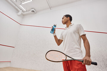 low angle, african american sportsman holding bottle of water and squash racquet after playing game