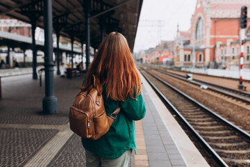 Back view of a young woman traveler with small backpack on the railway station, 30s women waiting for train. Tourism, solo vacation, rear view, no face - obrazy, fototapety, plakaty