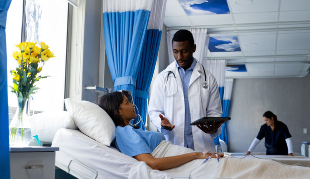 Diverse male doctor using tablet talking with female patient with iv drip in bed in hospital room